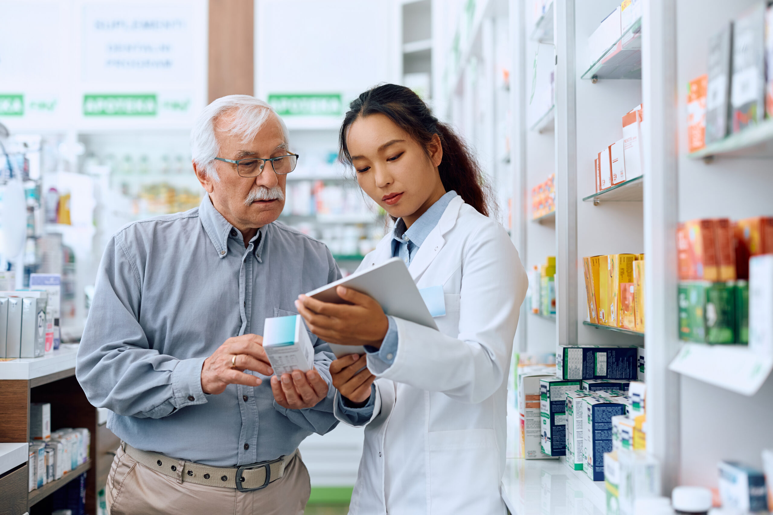 Female Chinese pharmacist using digital tablet while assisting senior man in buying medicine in a pharmacy.